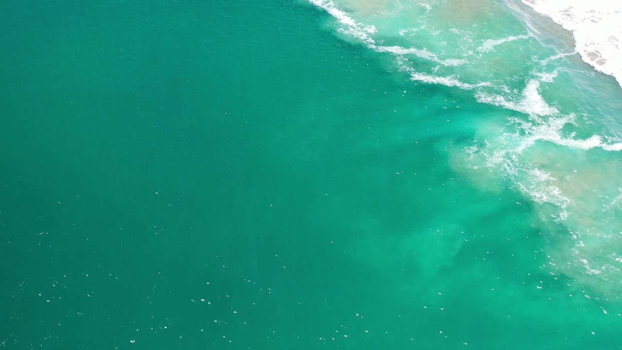 Overhead view of turquoise ocean water in Los Cabos, Mexico, with a band of white surf and pale shallow water at the upper right, no visible people.