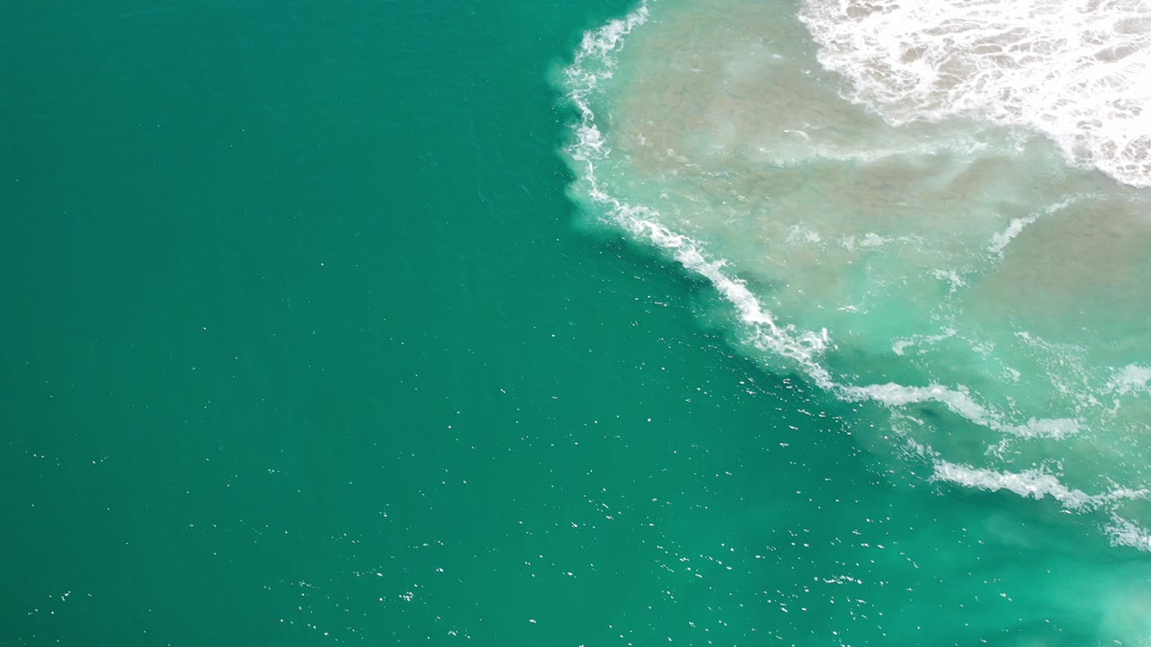 Overhead view of turquoise water and white shore foam in Los Cabos, Mexico, with the sandy shallows entering from the upper right and no visible people.