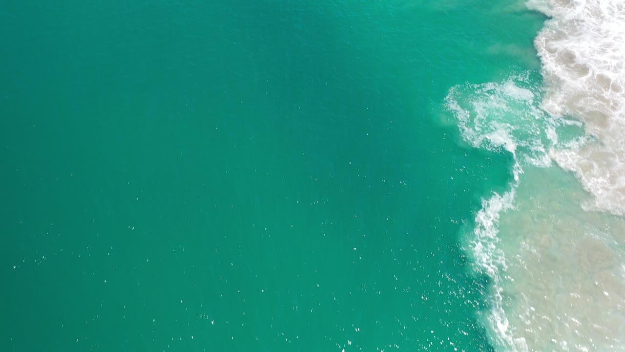 Overhead view of teal ocean water in Los Cabos with white foam breaking along the right edge and no visible people.