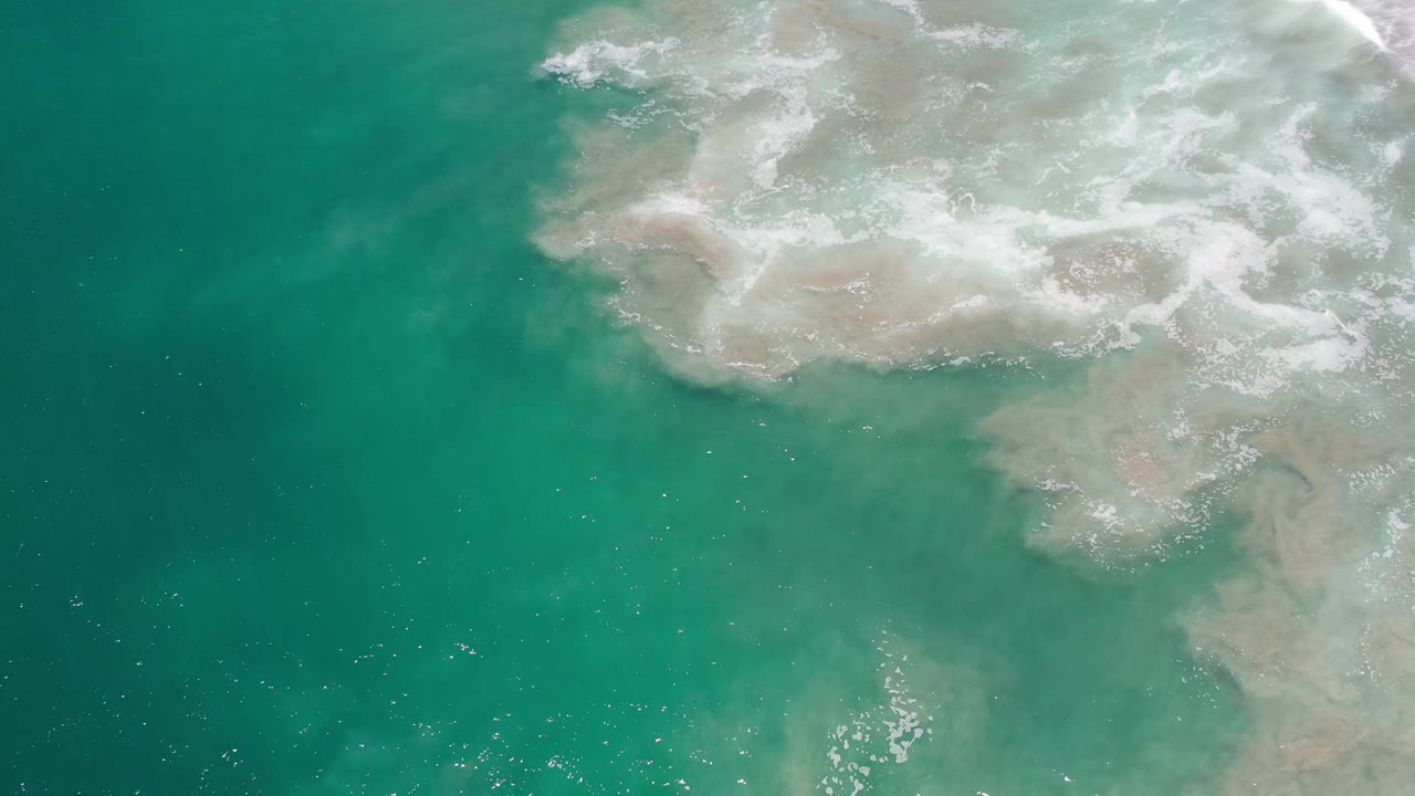 Overhead Los Cabos ocean frame with a broad white foam patch drifting across the top half and clear green water below, with no visible people.