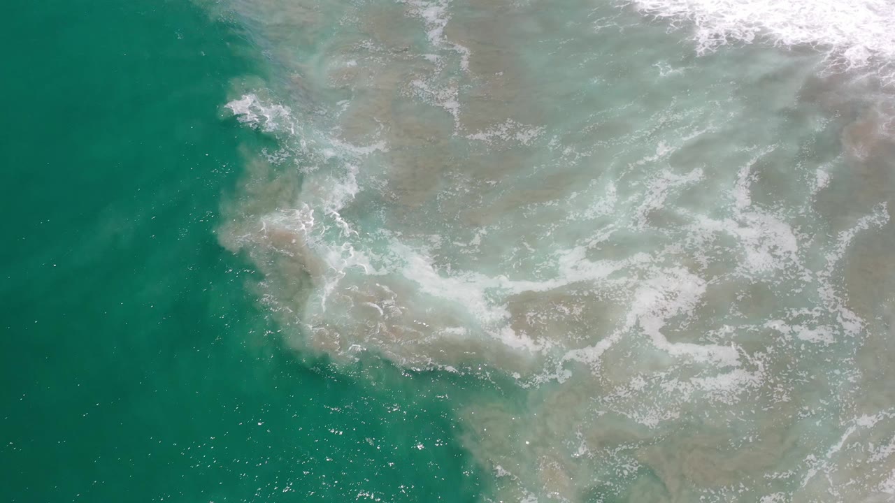 Aerial Los Cabos surf frame showing pale foam drifting across green water, with brown sand visible beneath and no visible people.