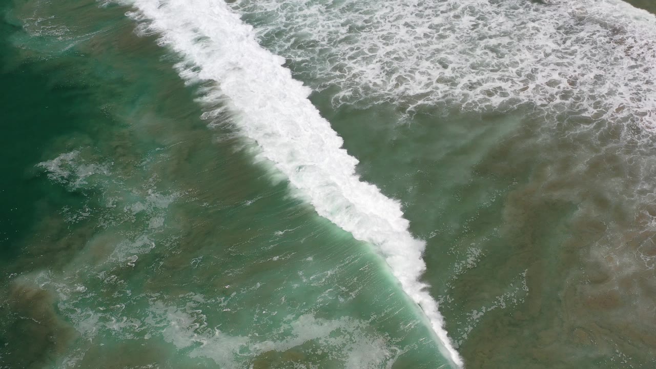 Top-down aerial view in Los Cabos, Mexico, with a narrow diagonal band of white surf over green and brown shallow water and no visible people.