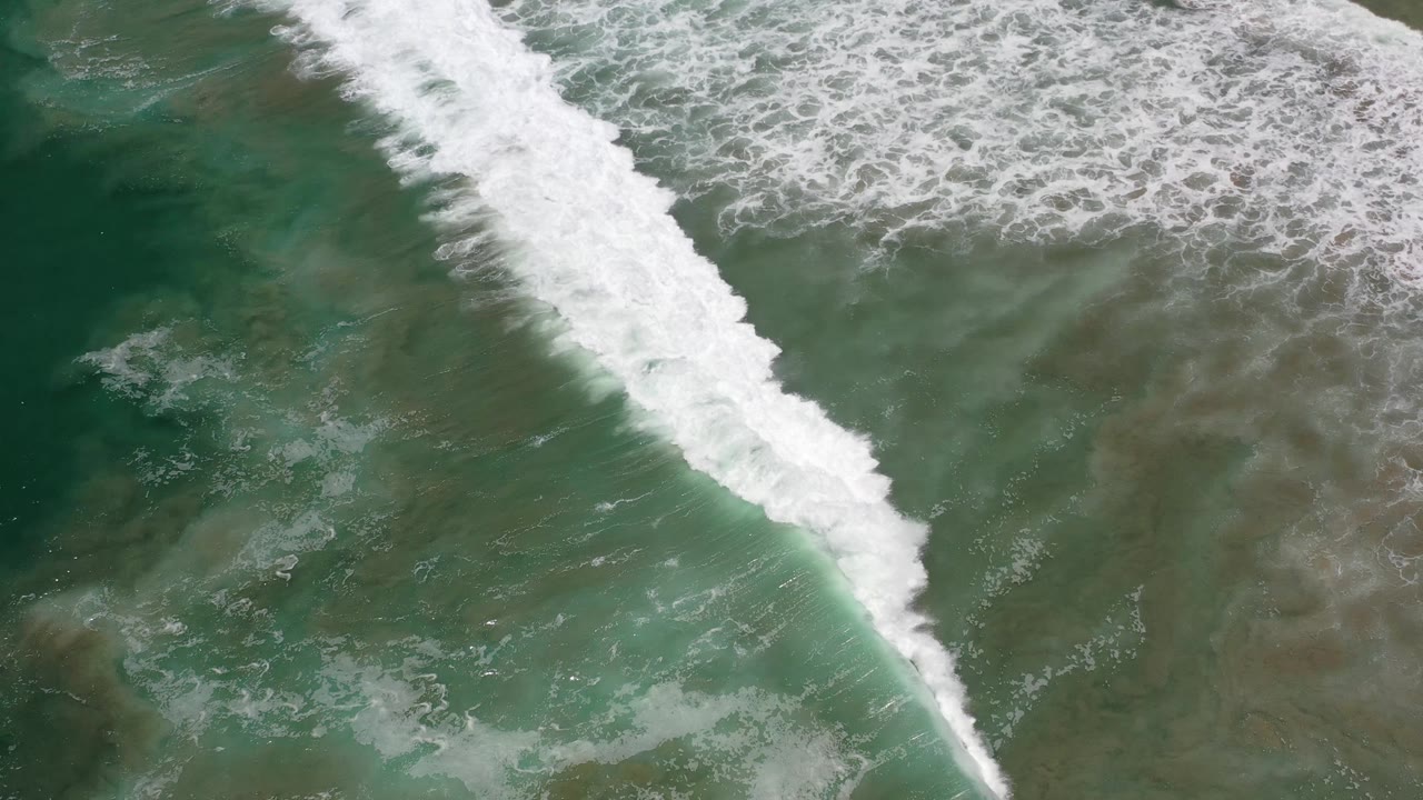 Aerial top view over Los Cabos water as a white wave band runs from upper left toward lower right across green shallows, with no visible people.
