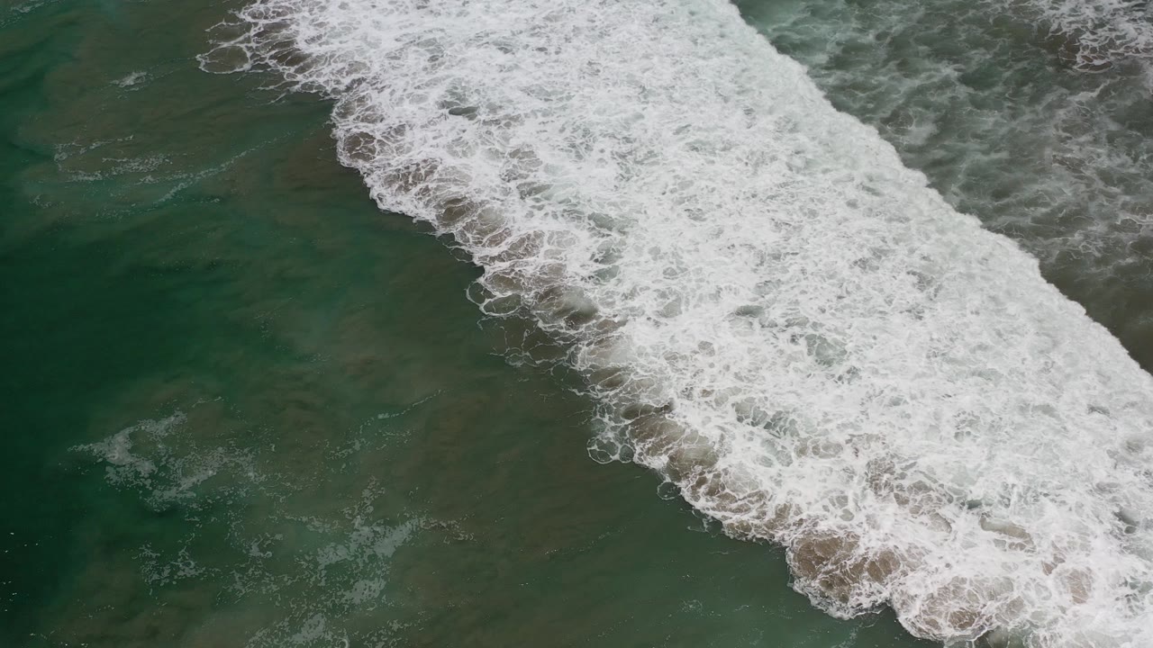 Overhead view of white surf breaking diagonally across green coastal water in Los Cabos, Mexico, with no visible people.
