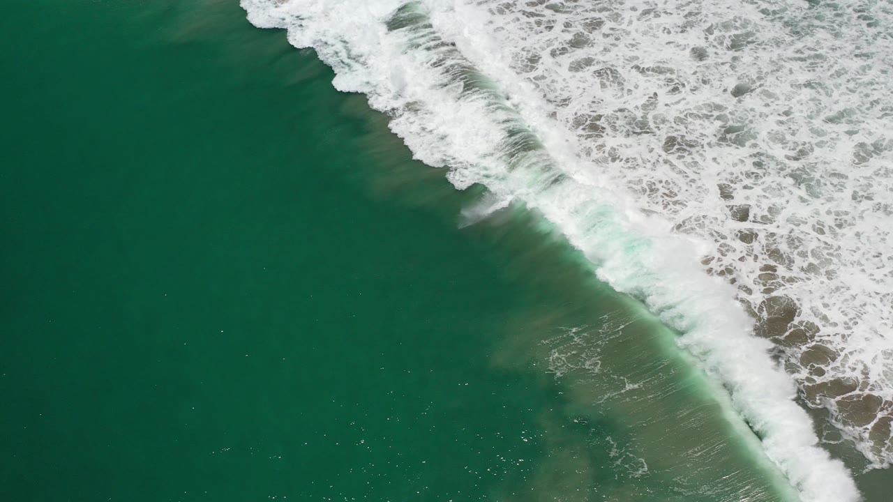 Overhead view of deep green Los Cabos sea water with a diagonal white foamy wave entering from the upper right, no visible people.