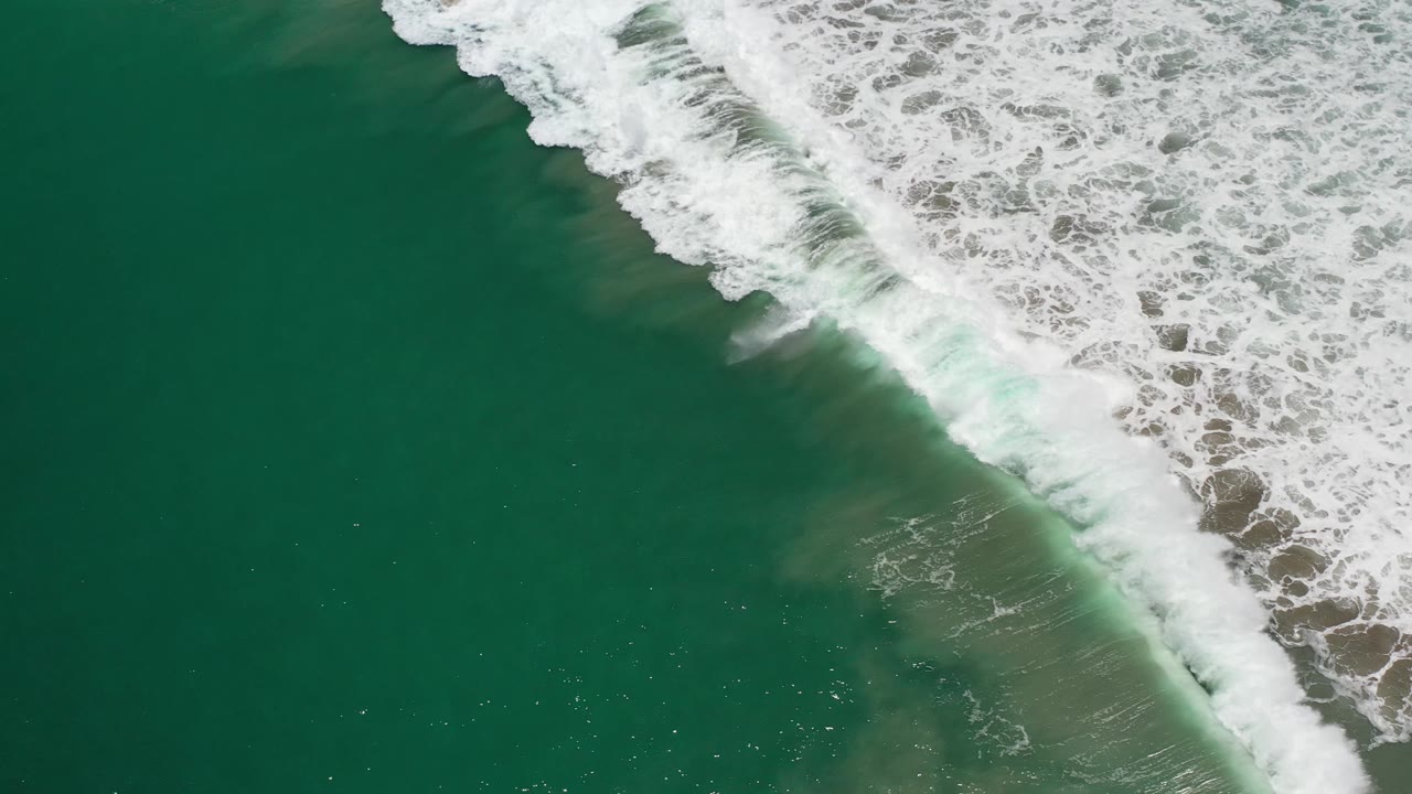 A broad white wave line breaks diagonally across green Los Cabos water, with darker open water on the left and no visible people.