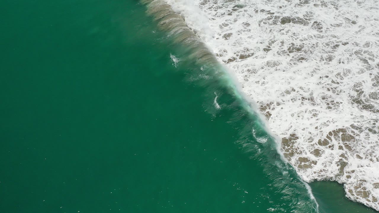 Overhead Los Cabos shoreline view with green water on the left, white surf breaking diagonally over wet sand on the right, and no visible people.