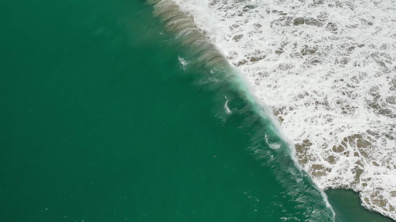 Aerial Los Cabos frame with a diagonal edge of white foam and tan wet sand against darker green ocean, with no visible people or structures.