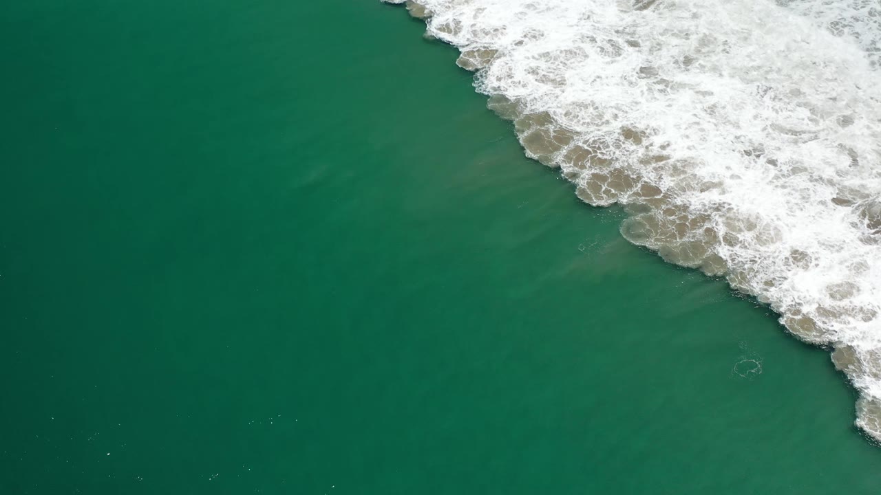 A diagonal strip of white wave foam pushes across green water at Los Cabos, Mexico, with open sea filling the left side and no visible people.