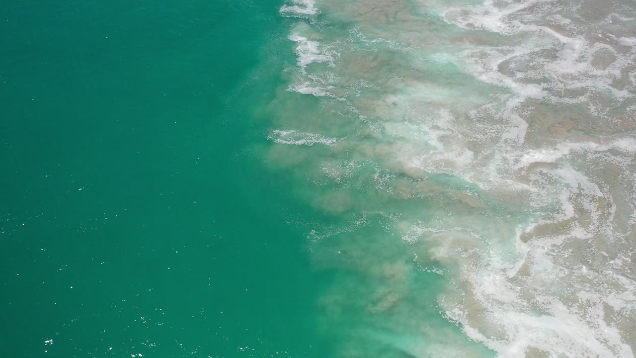 Overhead view of green ocean water in Los Cabos, Mexico, with white foam spreading over pale shallows on the right and no visible people.