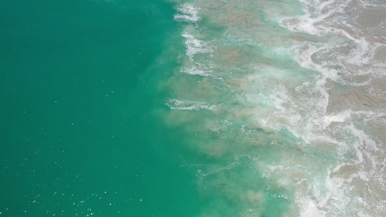 Overhead view of turquoise ocean water in Los Cabos, Mexico, with white foam crossing pale shallow areas on the right and no visible people.