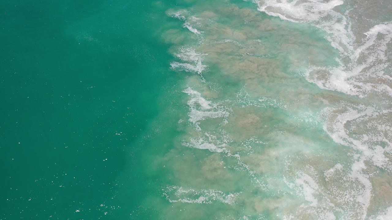 Overhead view of turquoise coastal water in Los Cabos, Mexico, with pale sand patterns below the surface, white foam along the right edge, and no visible people.