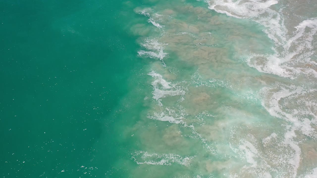 Aerial view of shallow Los Cabos ocean water with green-blue color, pale underwater sand patches, scattered bubbles, and no visible people.