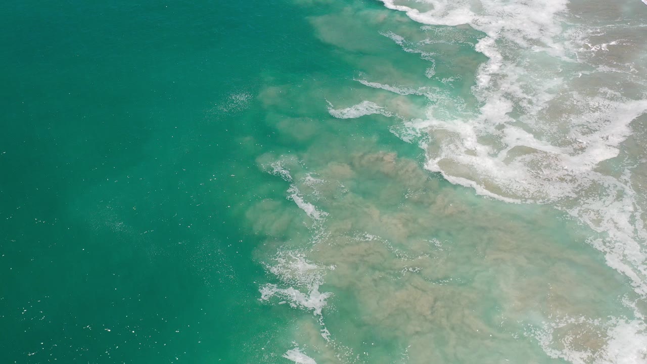 Overhead view of green water and pale submerged rocks in Los Cabos, Mexico, with white surf curling across the upper right and no visible people.