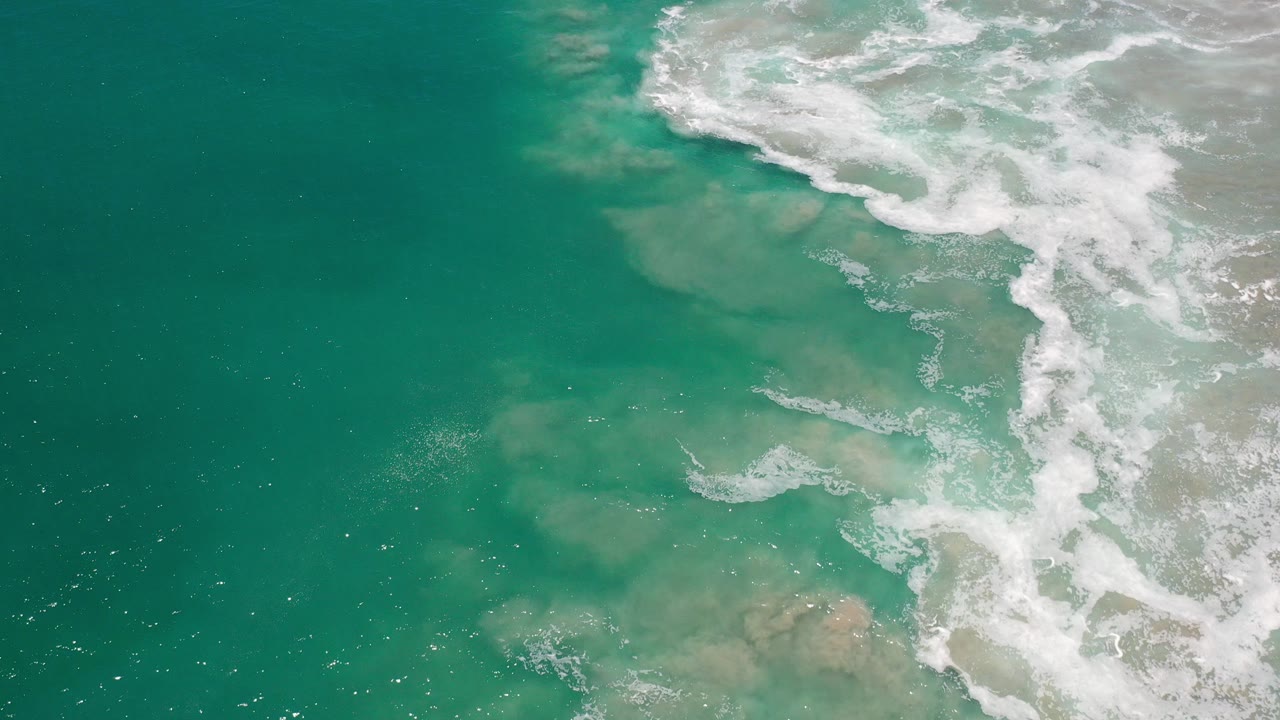 Overhead view of turquoise ocean in Los Cabos, Mexico, where white foam enters from the upper right over sandy shallows, with no visible people.