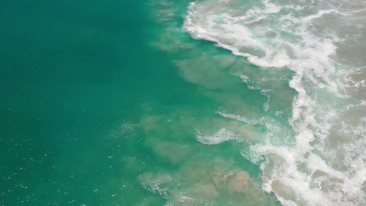 Aqua ocean surface in Los Cabos, Mexico, with a broad white breaker curling along the upper right and pale sand visible below the water, with no visible people.