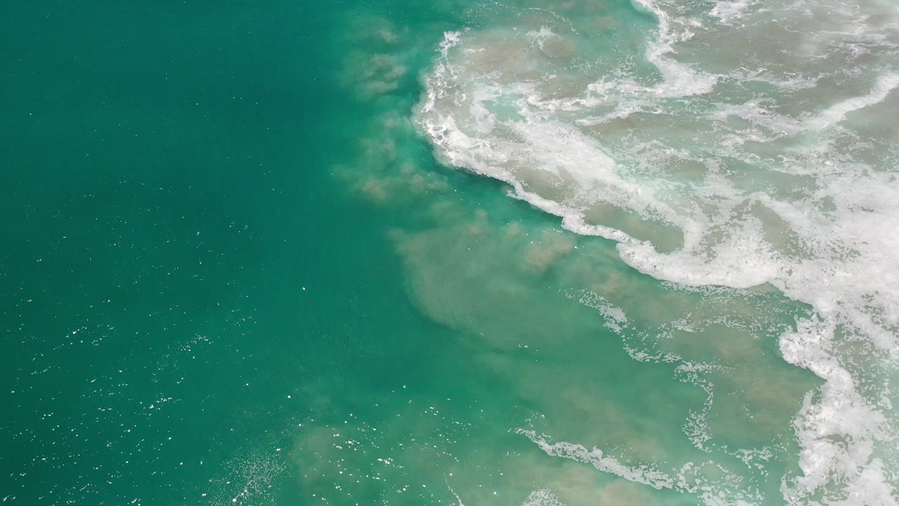 Overhead view of turquoise ocean and white surf crossing pale submerged sand in Los Cabos, Mexico, with no visible people.