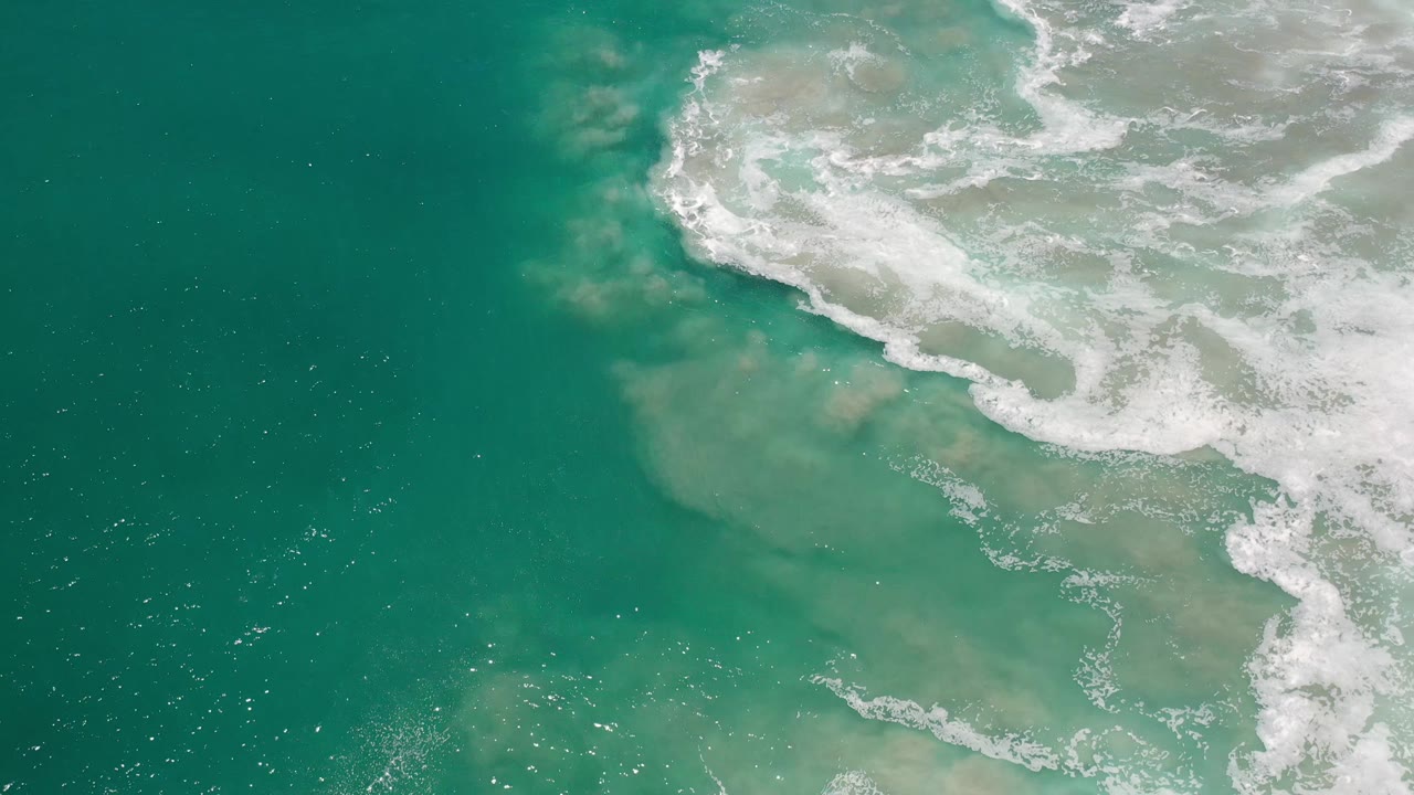 A diagonal band of white wave foam divides blue-green ocean from cloudy sand-toned shallows in Los Cabos, Mexico, with no visible people.