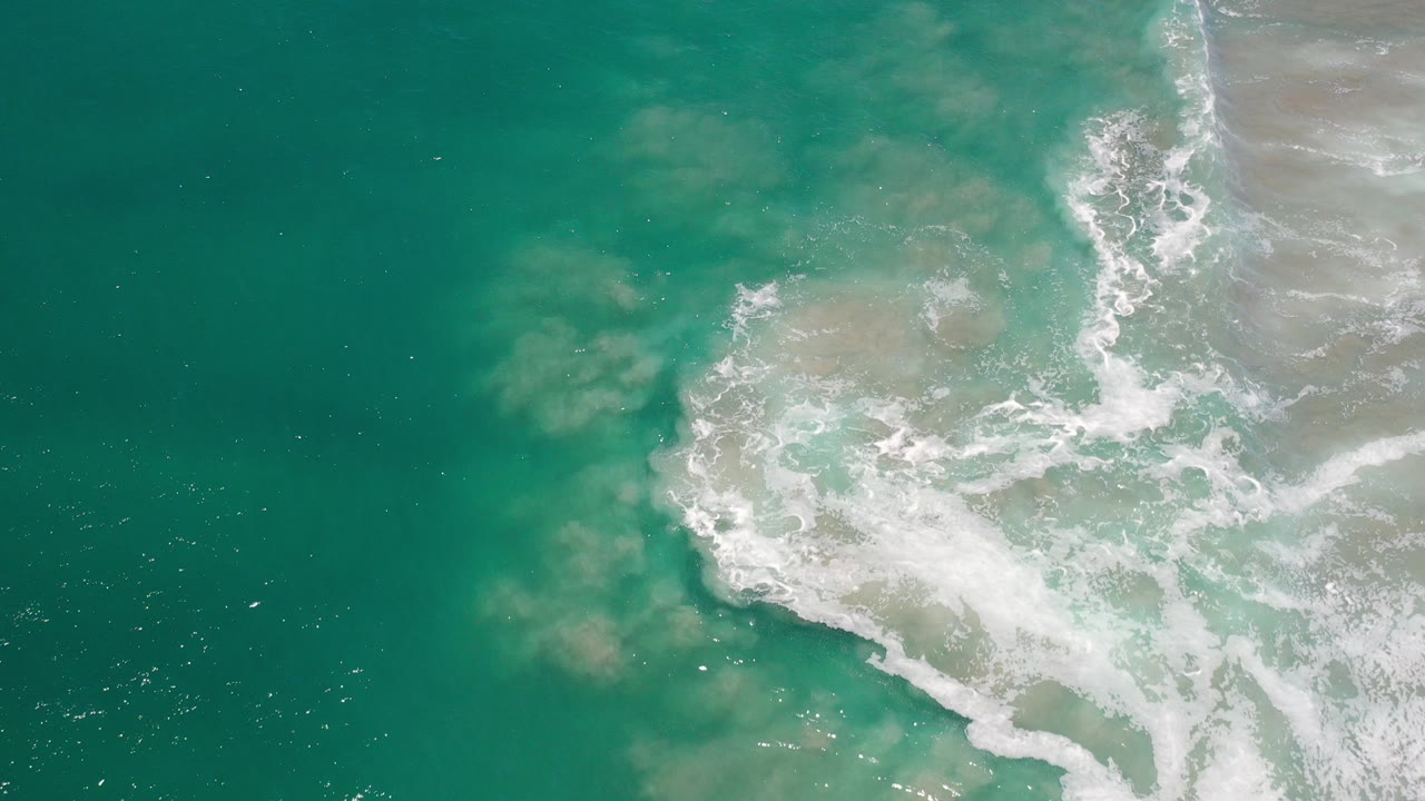 Top-down aerial frame from Los Cabos, Mexico, showing green-blue water, pale sandy patches, and white surf curling through the right half with no visible people.