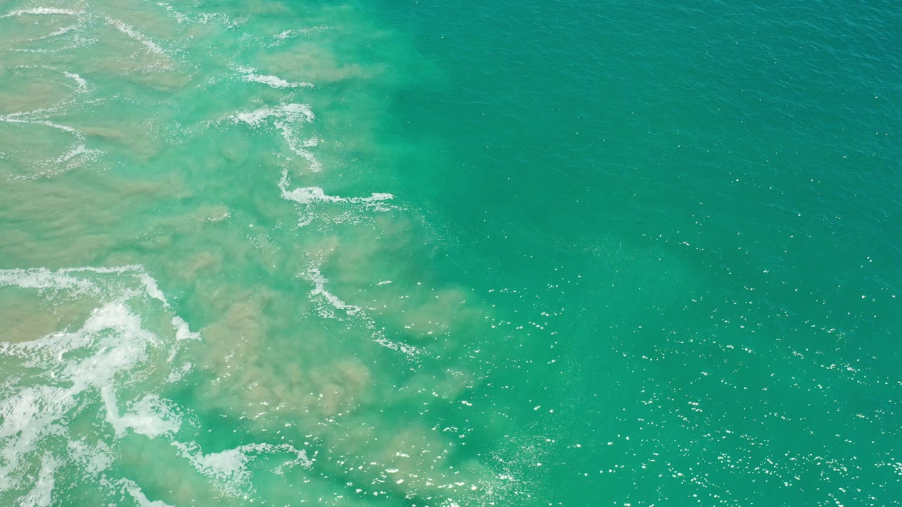 Overhead view of turquoise Los Cabos ocean water with pale sandbars under the surface, thin white foam along the left side, and no visible people.