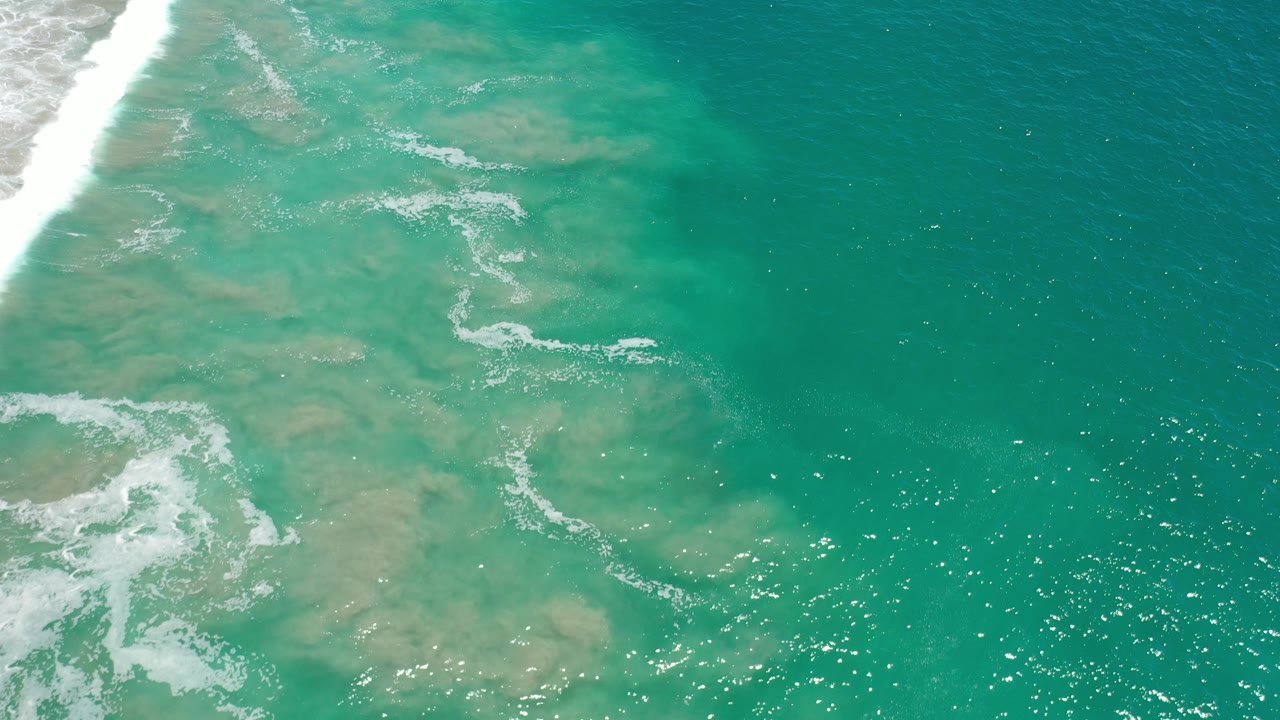 Aerial view of turquoise Los Cabos water with pale sandy patches, white surf entering from the left, sun sparkles on the surface, and no visible people.