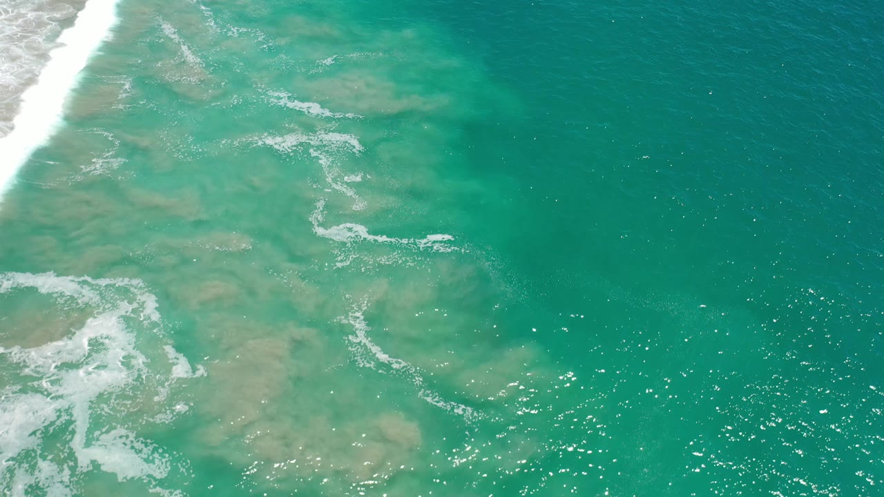 Aerial Los Cabos water frame with light tan shallows beneath the surface, curved foam streaks near the center, and no visible people.