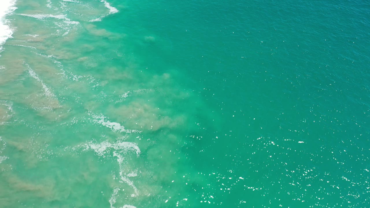 Overhead view of green-turquoise water in Los Cabos with pale sandy shallows at left, small white wave lines, sun sparkles, and no visible people.