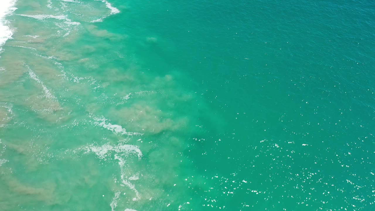 Overhead Los Cabos water view with milky sand patches on the left, darker green water to the right, scattered foam, and no visible people.