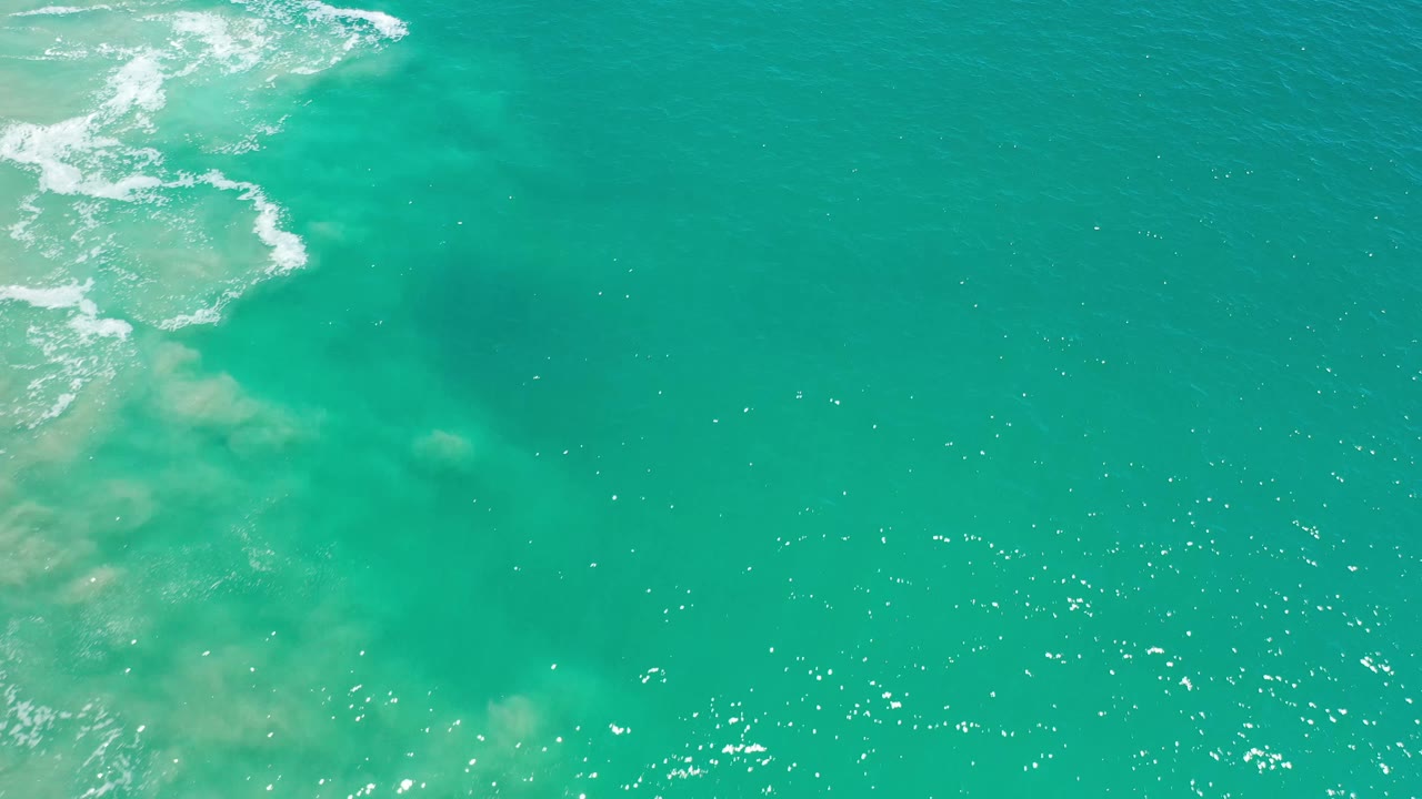 Aerial ocean frame in Los Cabos, Mexico, showing blue-green water, pale sandy shallows near the left side, scattered sun glints, and no visible people.