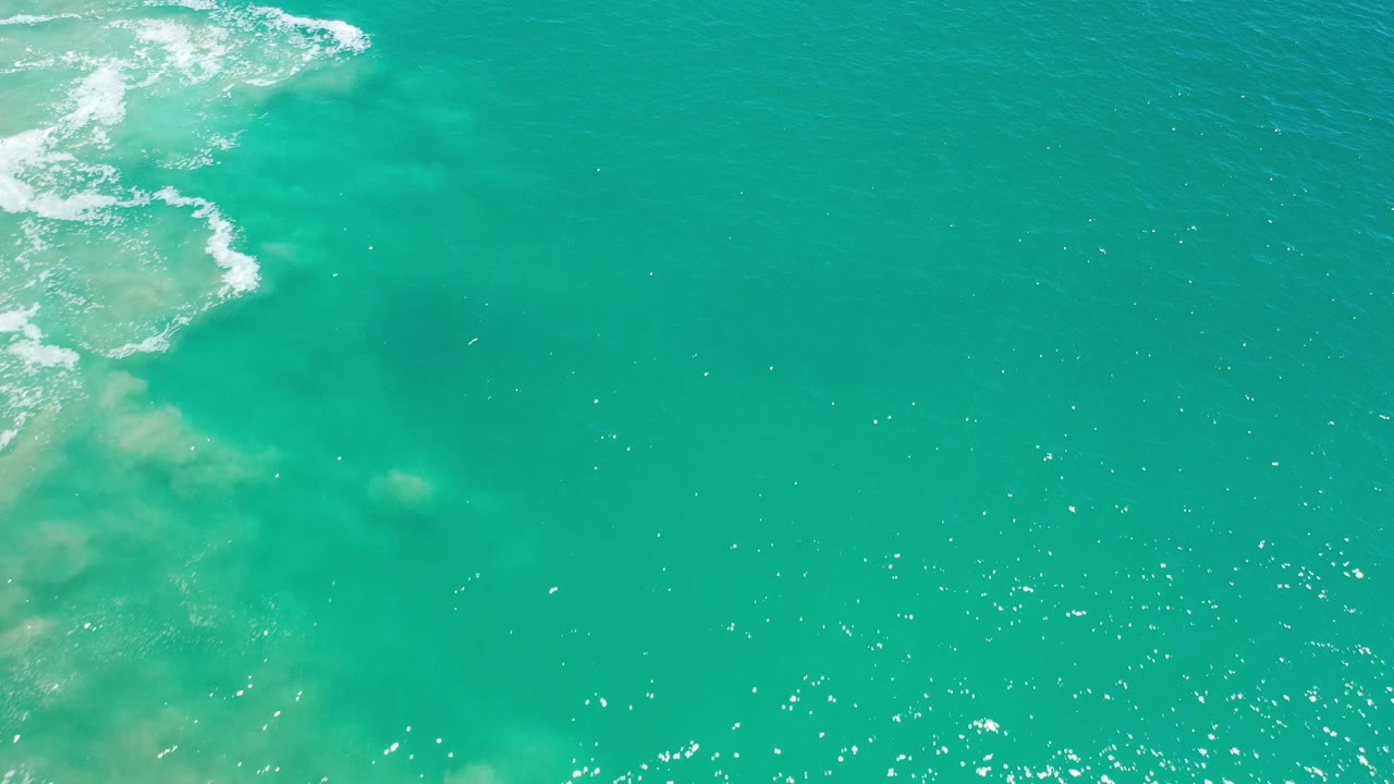 Open turquoise sea with white foam in the upper left and bright surface sparkle.