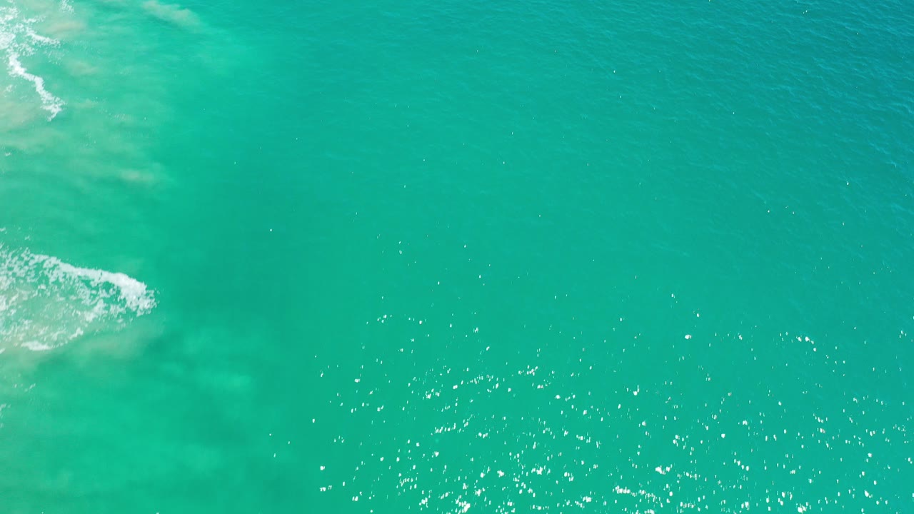Aerial view over turquoise green ocean in Los Cabos, Mexico, with pale surf entering from the upper left and no visible people.