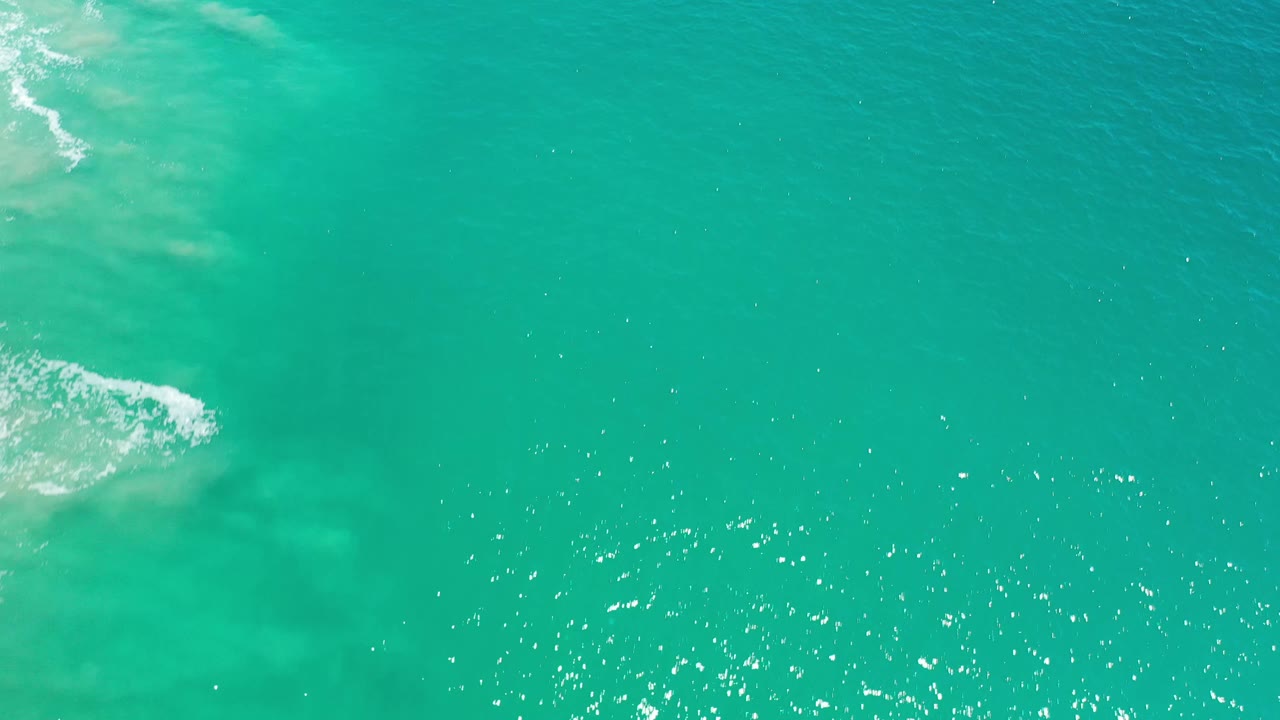 Aerial frame of green-blue Los Cabos ocean with scattered sun sparkle and a thin patch of white surf along the left edge, no visible people.