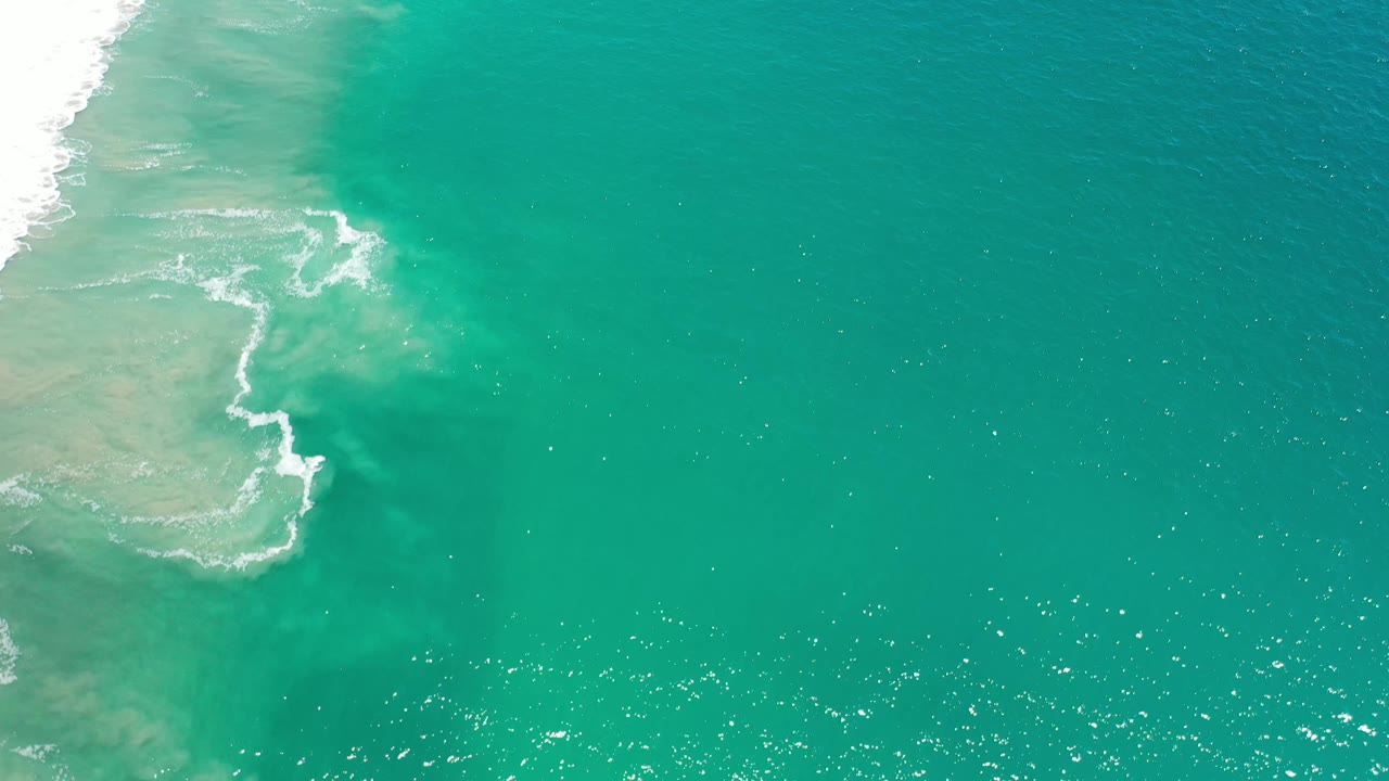 Blue-green ocean fills most of the frame while a curved band of white foam traces the left shoreline in Los Cabos, Mexico, with no visible people.