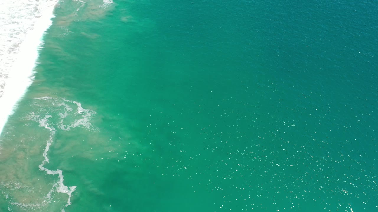 Overhead view of green ocean water in Los Cabos, Mexico, with a white surf band along the left edge, pale underwater tones, sun flecks, and no visible people.