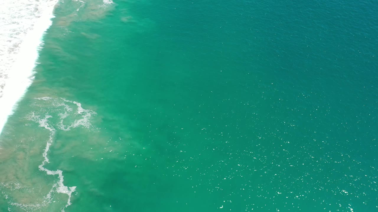 Aerial frame over green sea in Los Cabos, Mexico, where white foam curls near the left side and small bright reflections scatter across the open water, with no visible people.