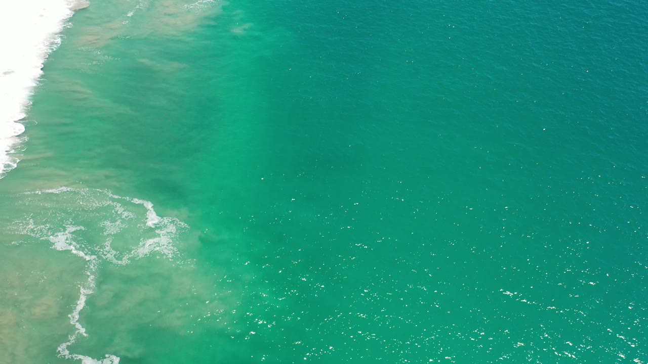 Aerial view over Los Cabos, Mexico, with white surf entering from the left, pale sand visible below turquoise water, and no visible people.