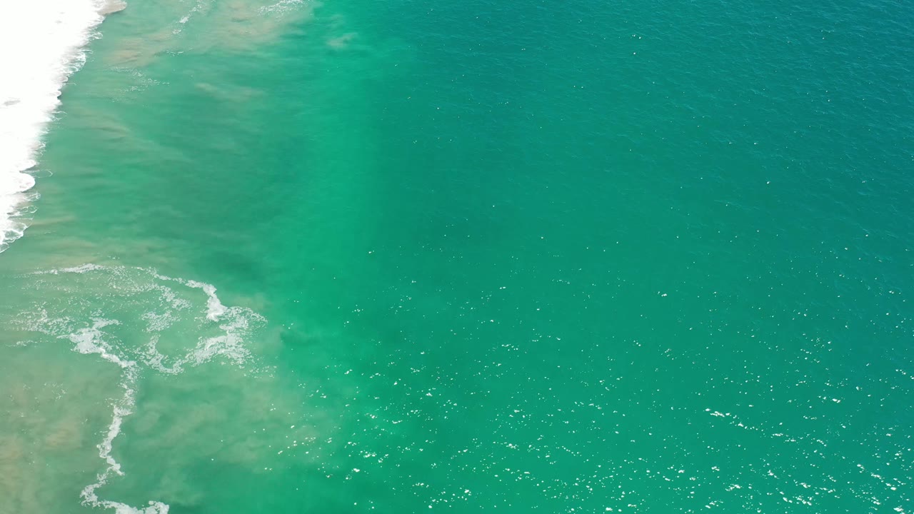 Aerial Los Cabos frame with a patch of white foam curling near the left side, deeper teal water to the right, and no visible people.