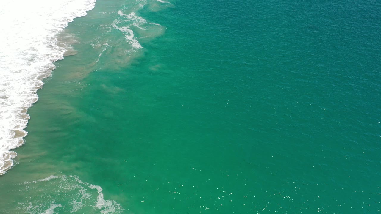 Overhead view of green-tinted ocean water in Los Cabos, Mexico, with white surf entering from the upper left and no visible people.
