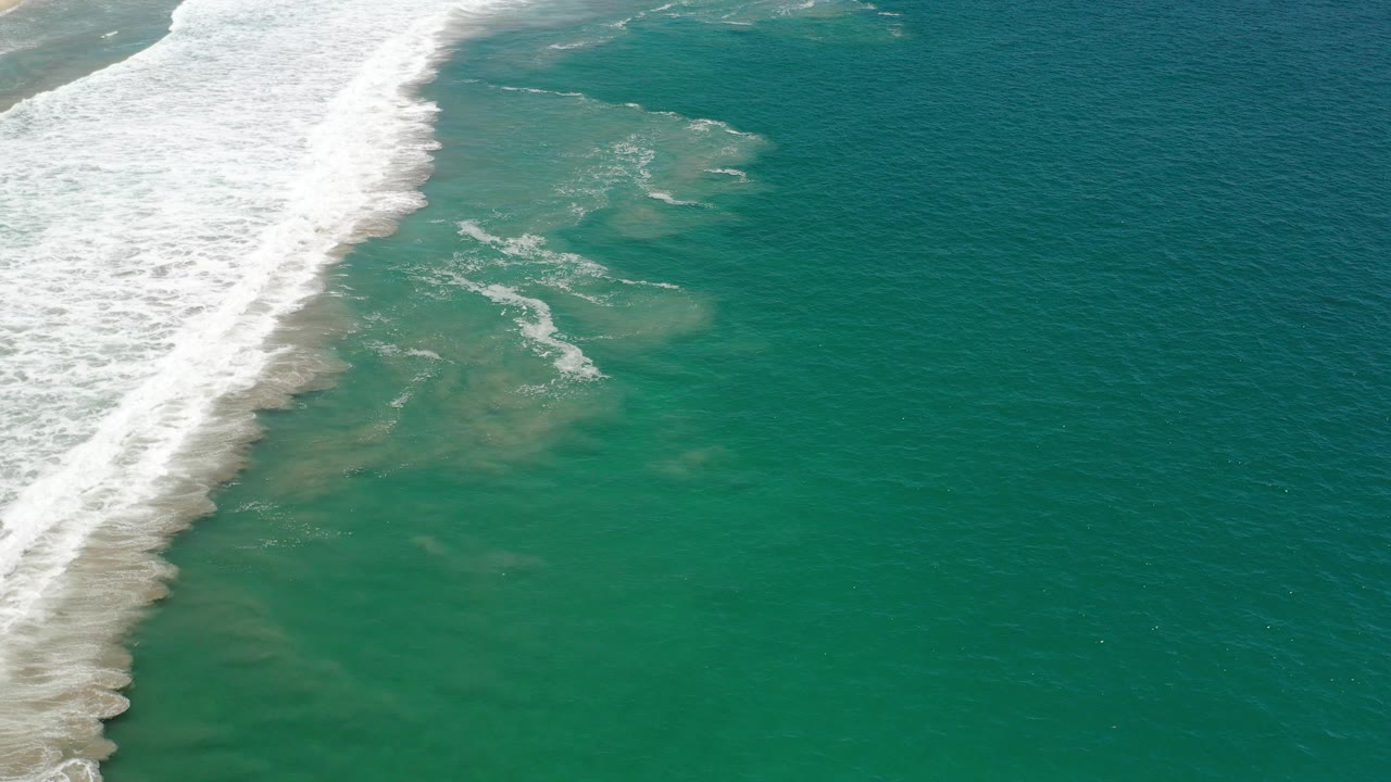 Aerial frame from Los Cabos, Mexico showing a diagonal band of white surf on the left and green ocean shallows across the center, with no visible people.