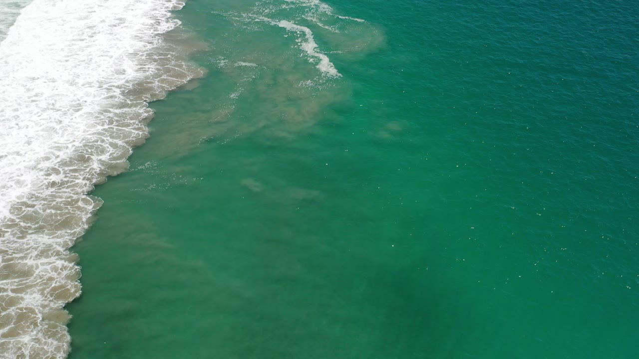 Aerial view over Los Cabos green ocean water with white surf pushing in from the upper left and no visible people.