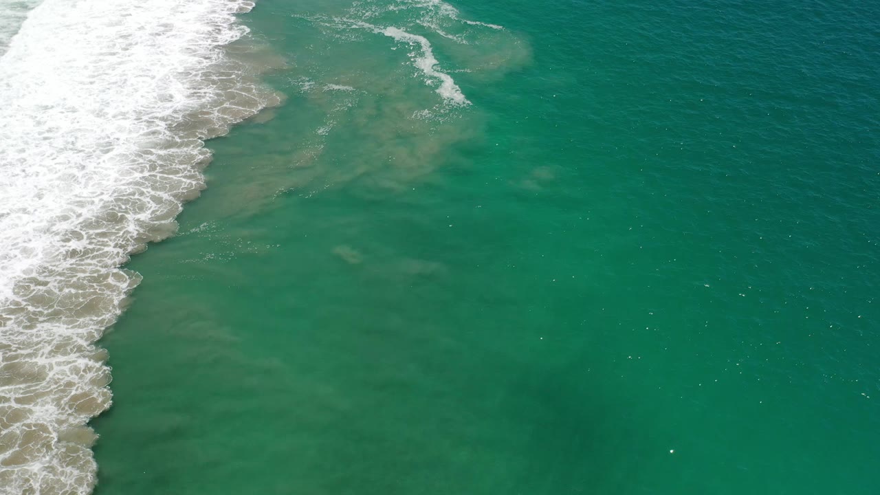 Aerial Los Cabos frame with foamy surf curving down the left side and clear green water spreading across the right, with no visible people.