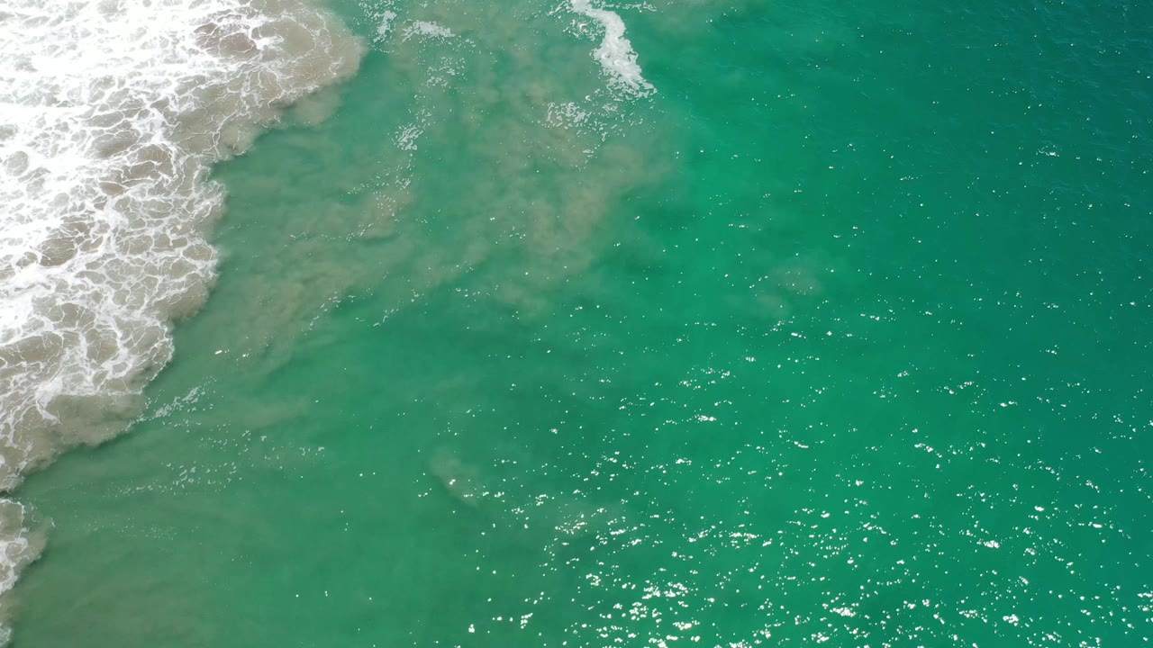 White foam expands farther along the left edge of the Los Cabos shoreline water while tan sand remains visible beneath the clear turquoise surface, with no visible people.