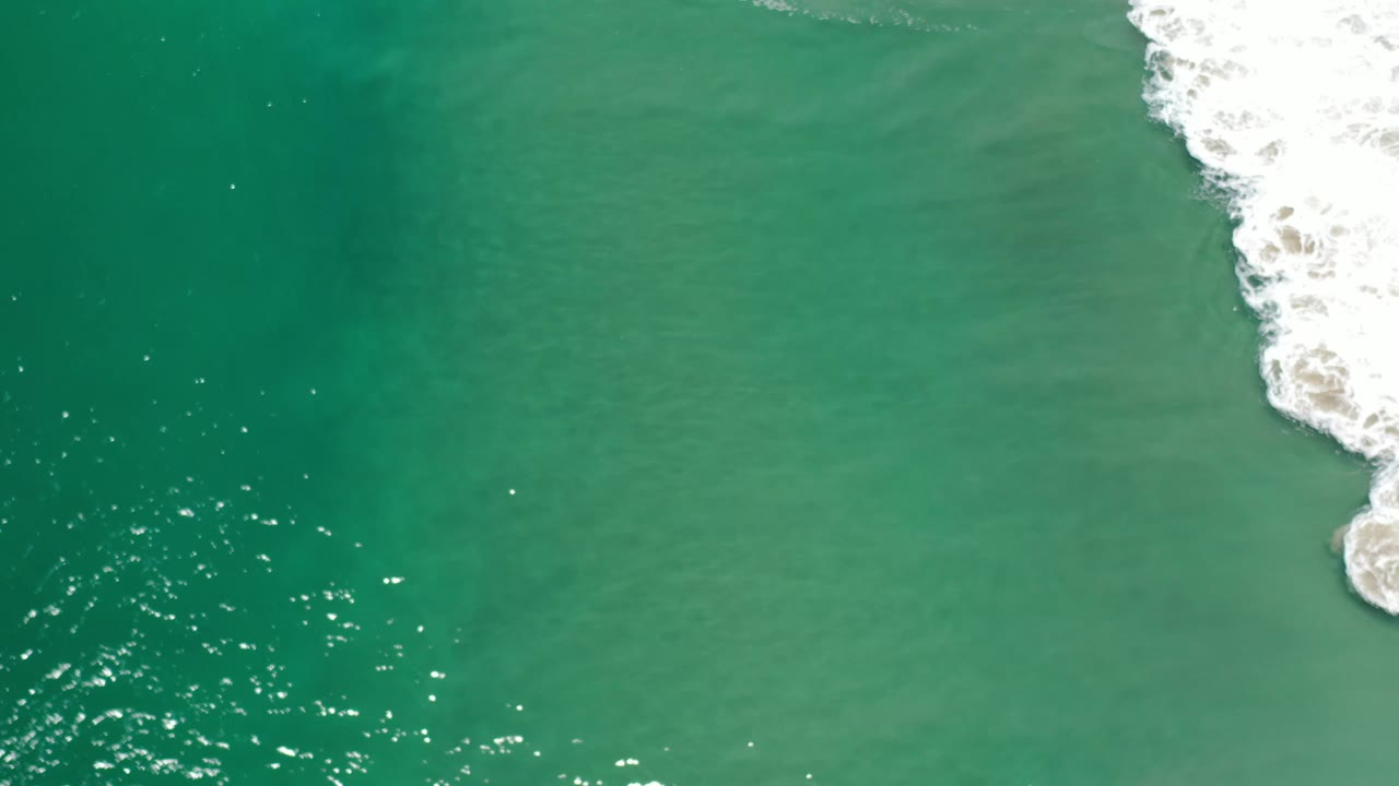 Teal sea surface in Los Cabos, Mexico, seen from above, with scattered sun glints across the lower left and a broken white wave line on the right, no visible people.