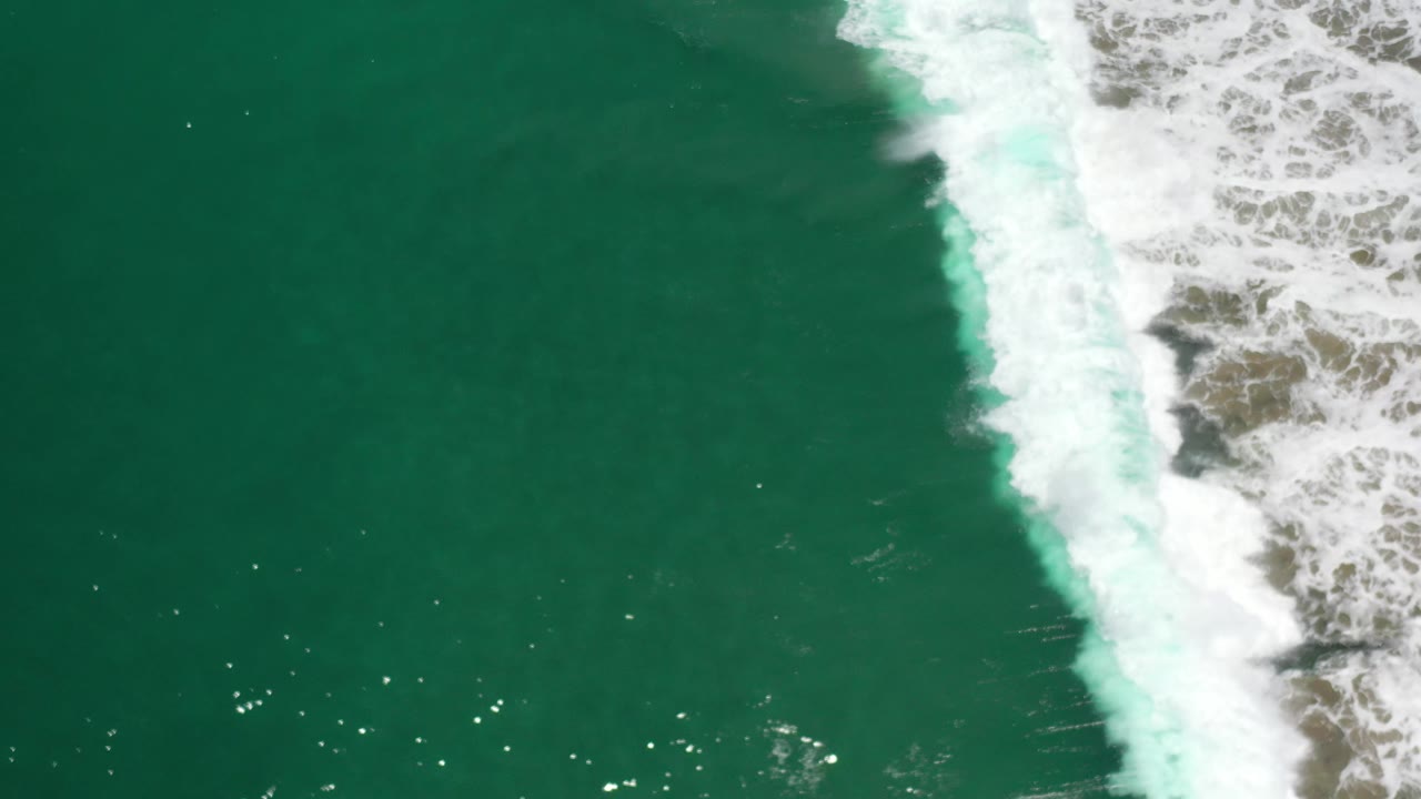 Overhead Los Cabos coastline frame with open green water on the left, white wave foam on the right, and no visible people.