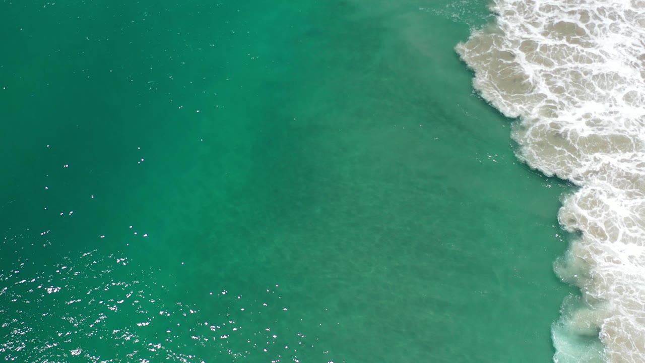 Overhead view of teal shallow water in Los Cabos, Mexico, with white foam breaking along the upper-right edge and no visible people.