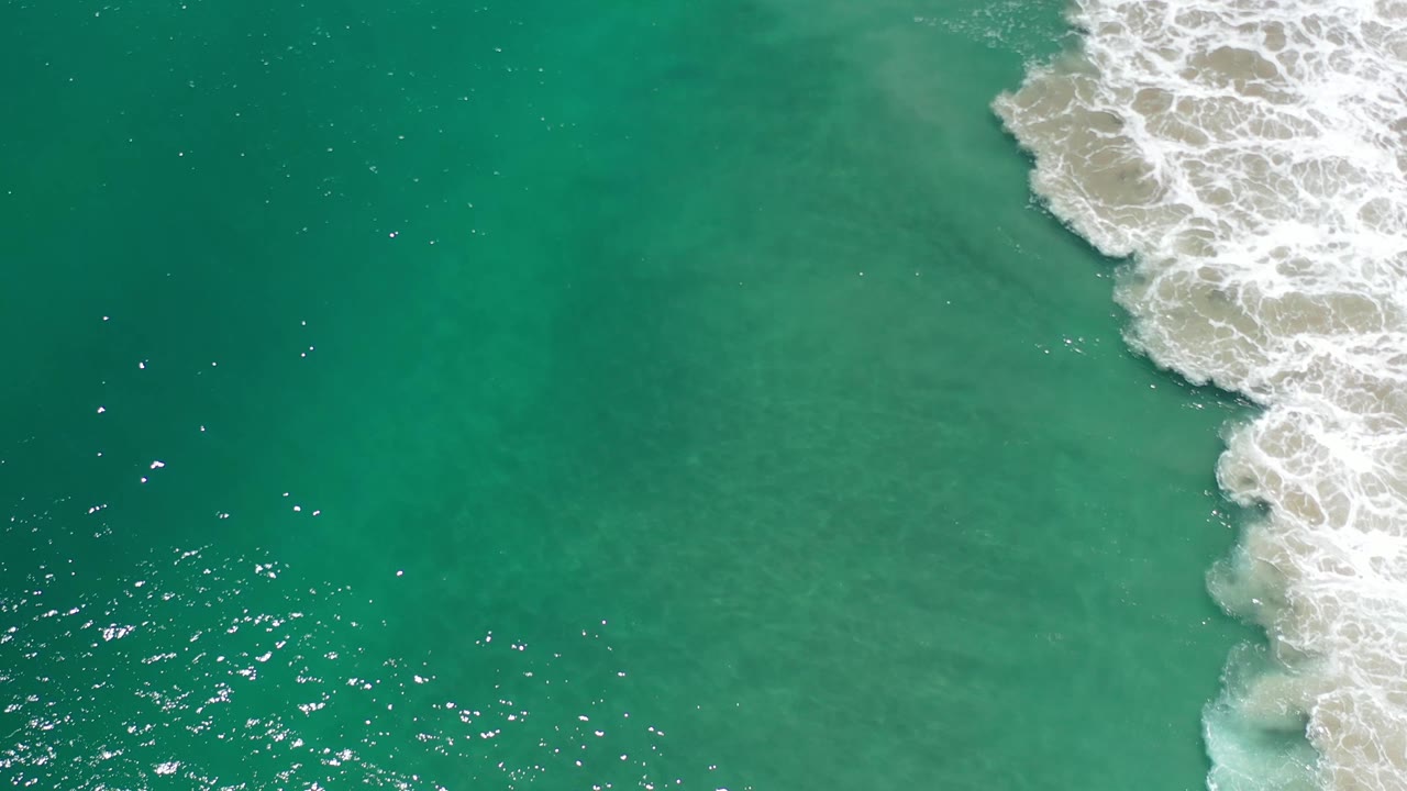 Overhead Los Cabos water frame showing green-blue shallows, a brighter patch of surf foam on the right, scattered sun reflections, and no visible people.