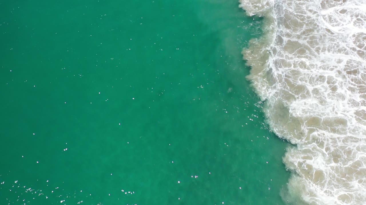 Overhead view of turquoise water in Los Cabos, Mexico, with white foamy surf entering from the upper right and no visible people.