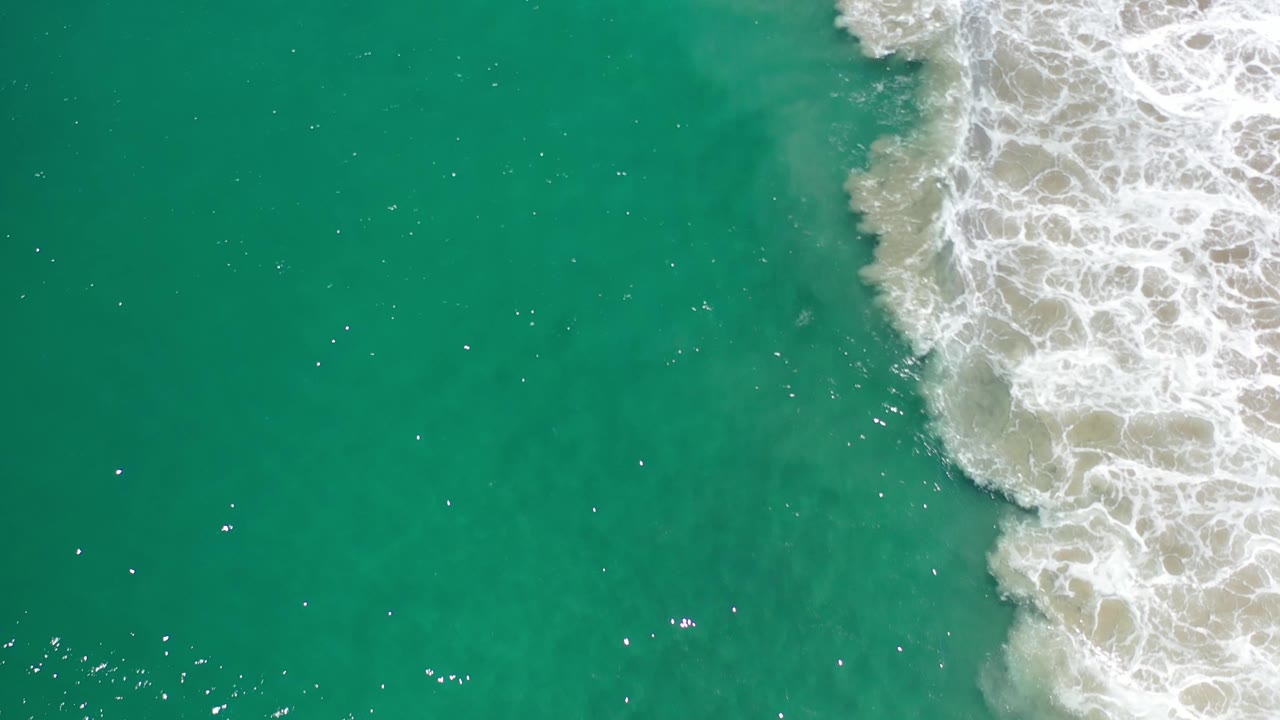 Aerial frame over Los Cabos aqua sea where the white foam line pushes farther into the right side of the water, with no visible people.