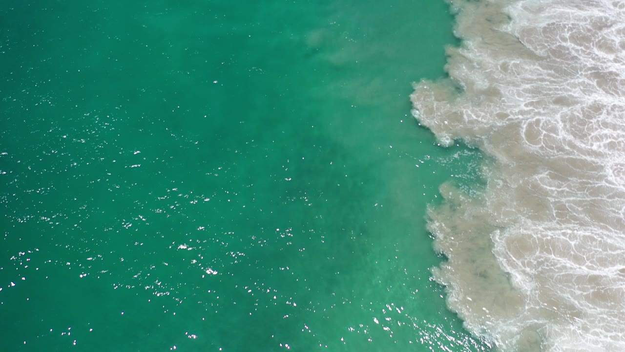Overhead view of turquoise ocean in Los Cabos, Mexico with sun glints on the water and white surf entering along the right edge, no visible people.