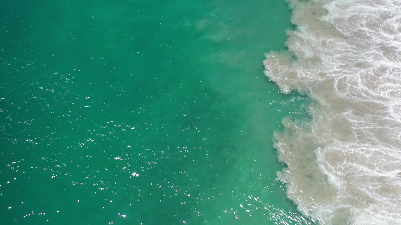 Aerial Los Cabos ocean frame with bright specks of reflected sunlight across turquoise water and a vertical band of white surf at the right, no visible people.